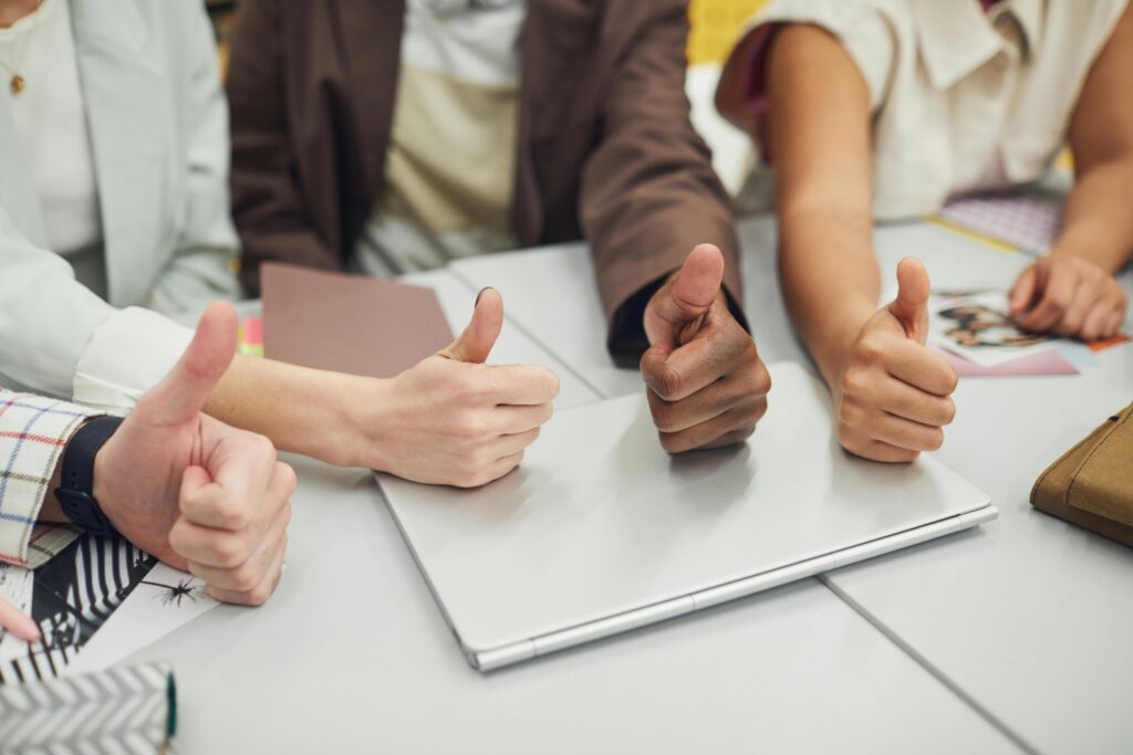 Four diverse hands showing thumbs up over a laptop on a desk, symbolizing teamwork and approval.